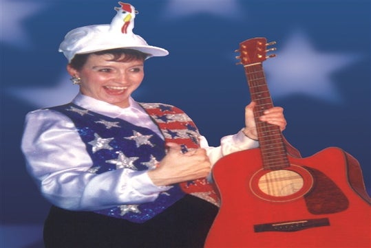 Patty Waszak in an American Flag vest and a white hat giving a thumbs up and holding a red guitar.