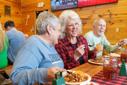 Three people smiling and eating barbecue meals at a wooden table in a rustic setting.