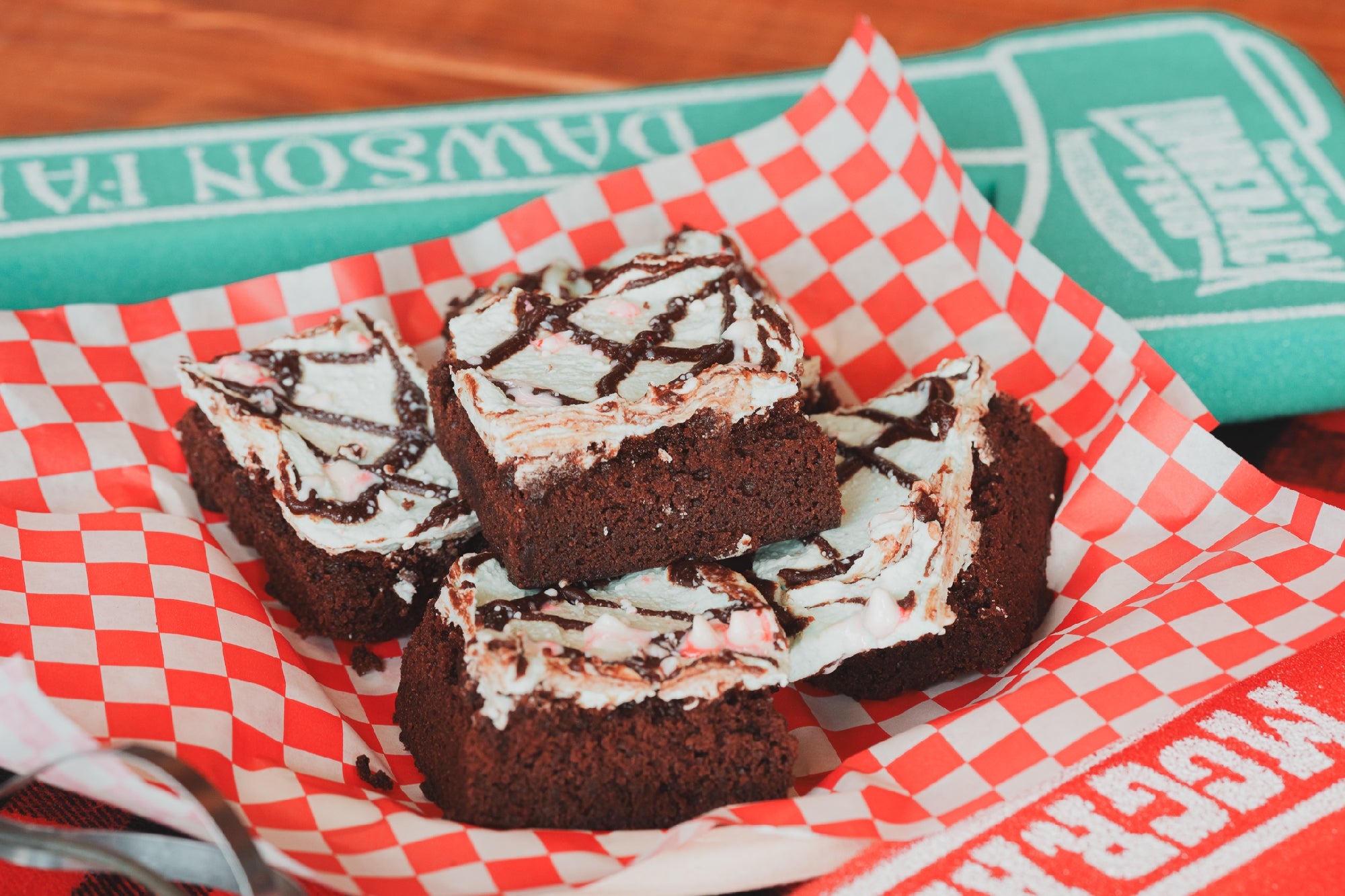 Four pieces of frosted brownies with chocolate drizzle are arranged on red and white checkered paper in a basket, with a green foam finger in the background.