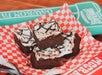 Four frosted brownies with chocolate drizzle on a red checkered paper, placed on a table near a green sports foam finger.