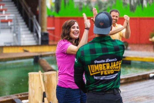 A woman high-fives a man in a "Lumberjack Feud" shirt outdoors, while another man claps in the background near a log pool.