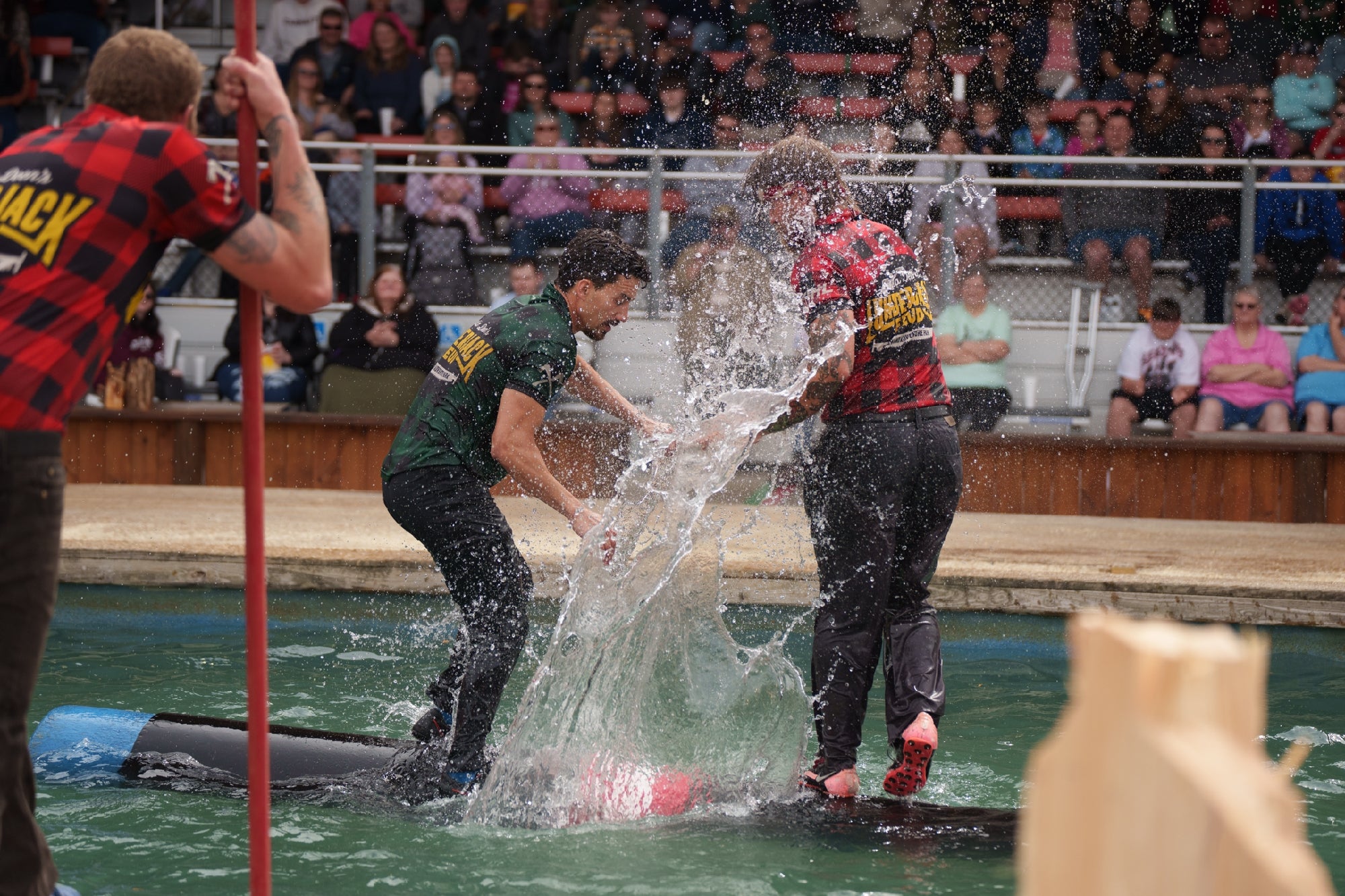 Two men wearing team shirts balance on a log over water, with one splashing the other. Spectators watch in the background.