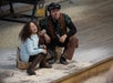 A person and a child sit on wooden blocks, sticking out their tongues playfully. Sawdust covers the wooden floor around them.