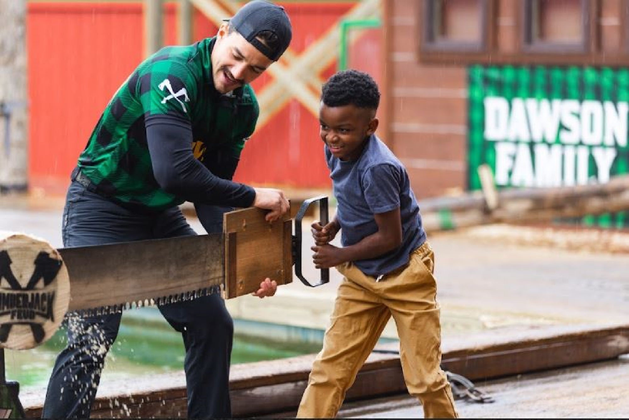 Man and child use a large saw together outdoors, smiling and engaged in a wood-cutting activity.