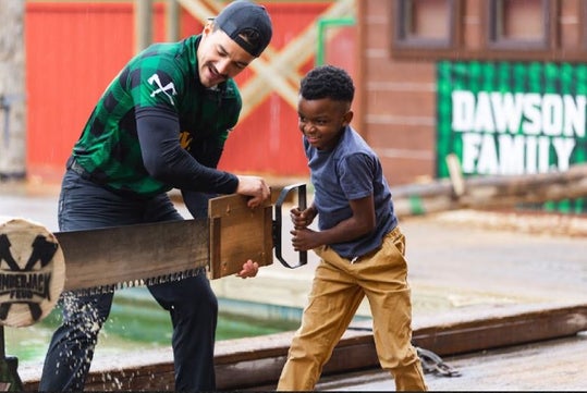A man and a boy use a large two-person saw to cut through a log outdoors, both focused and exerting effort. A sign in the background reads "Dawson Family.