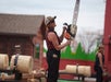 A person wearing a cap, sunglasses, and plaid shirt holds a chainsaw while standing next to logs on sawhorses at an outdoor lumberjack event.