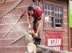 A lumberjack in a red plaid shirt standing on a log white also sawing on that same log with wood chips flying everywhere.