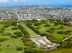 Aerial view of a cemetery with symmetrical rows of trees and graves, surrounded by green lawns, with a cityscape and ocean in the background.