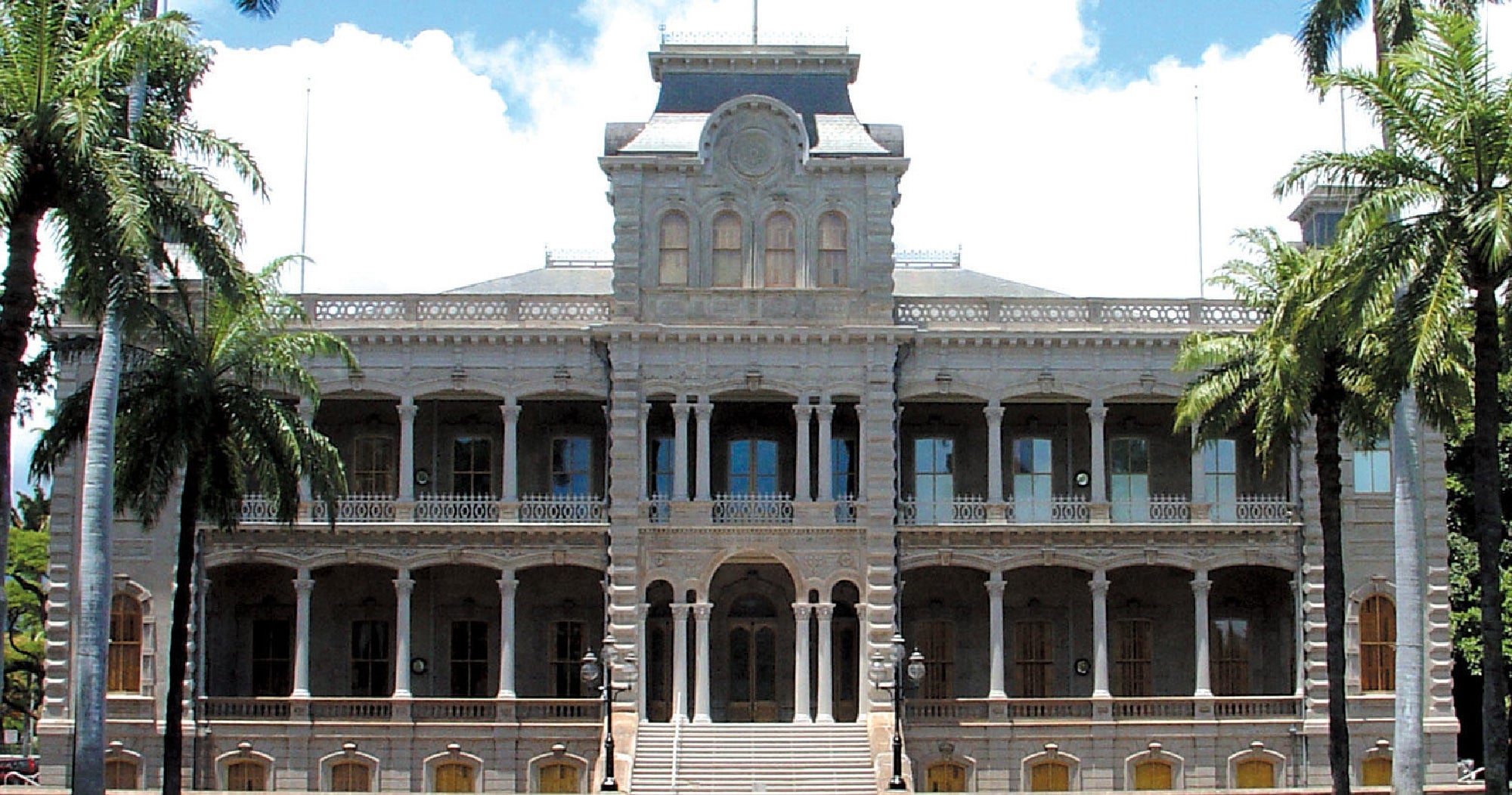 A large historic building with ornate architecture, arched windows, and two levels of columned balconies, framed by palm trees.