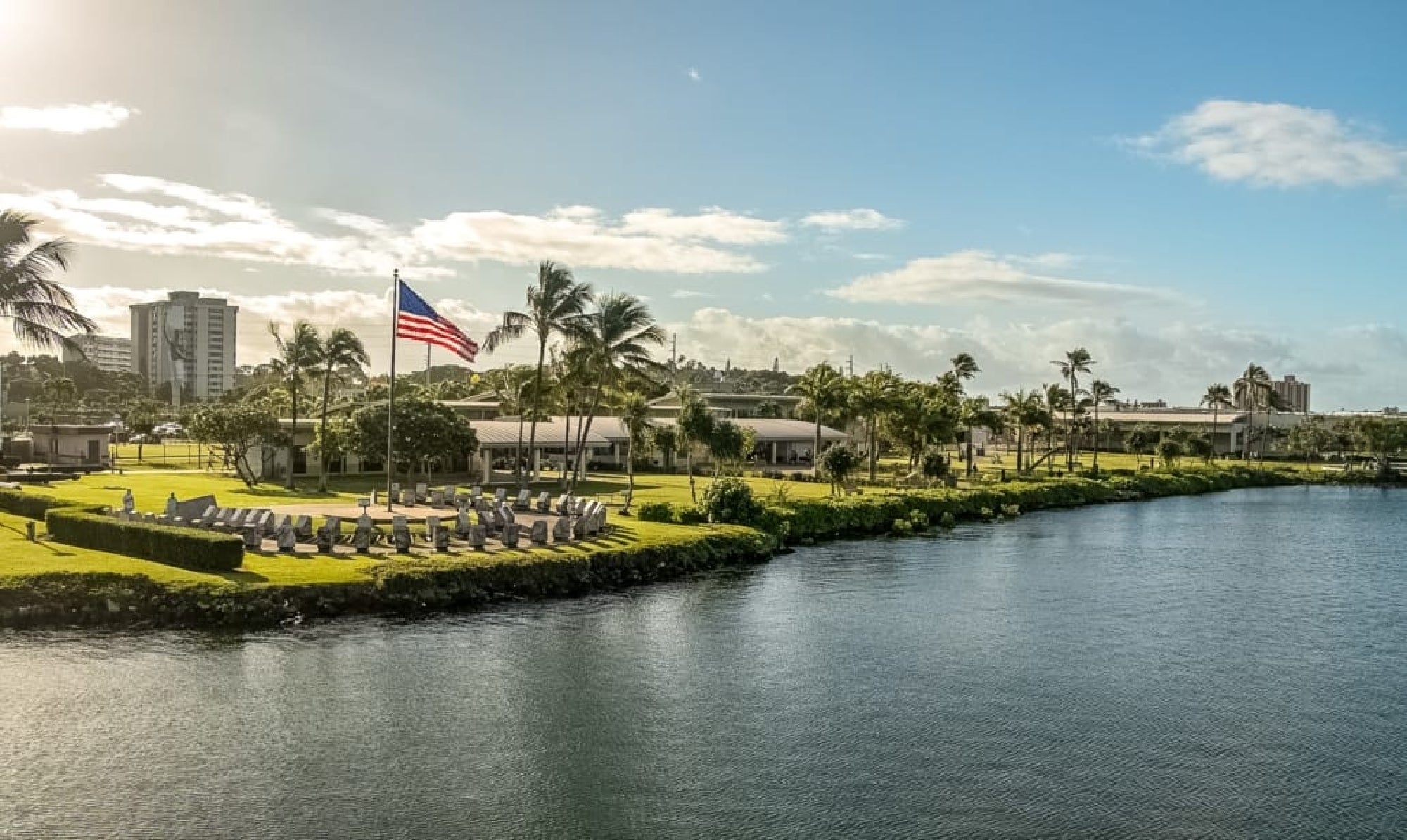A large American flag flies near a group of outdoor chairs on a grassy area by the water, with palm trees and buildings in the background under a partly cloudy sky.