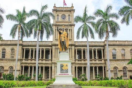 Statue of King Kamehameha I in front of Ali?iolani Hale, with palm trees and a clock tower in the background.