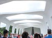 A crowd of people inside a white, open-air structure with geometric arches, looking towards a wall at the end.
