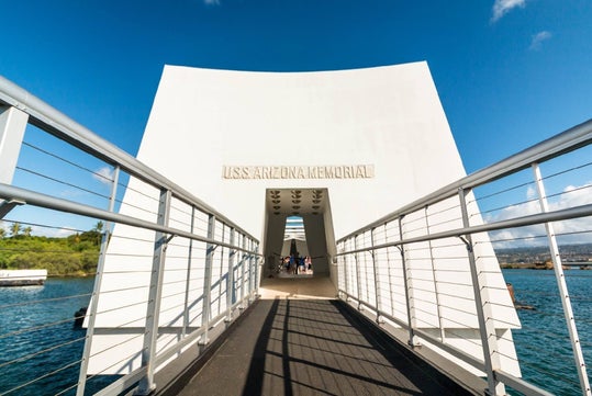 Walkway leading to the USS Arizona Memorial, a white structure over water, under a clear blue sky.