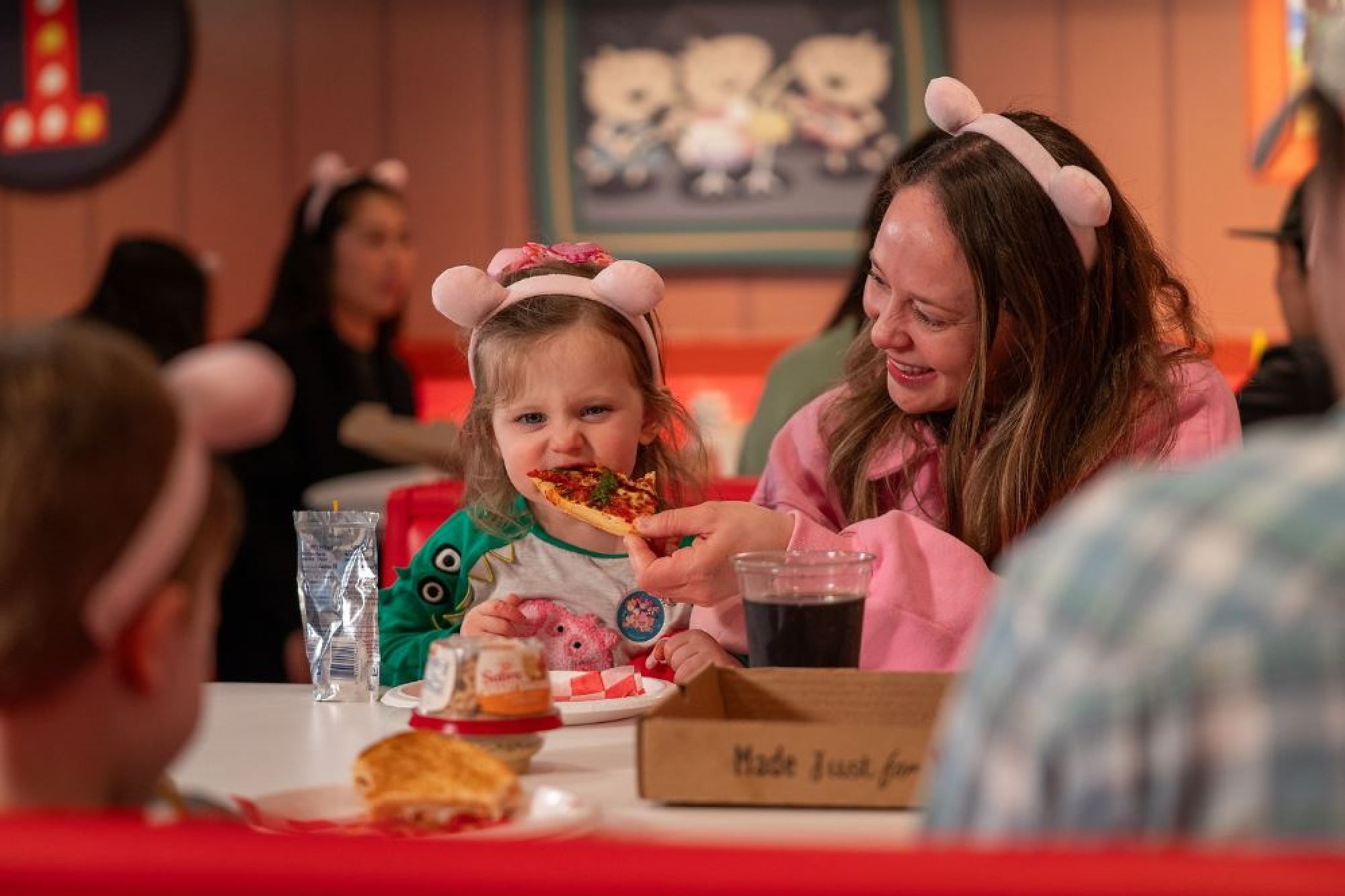 A woman and child wearing headbands sit at a table in a restaurant, sharing pizza and smiling. A pizza box and drink are on the table. Other people are in the background.