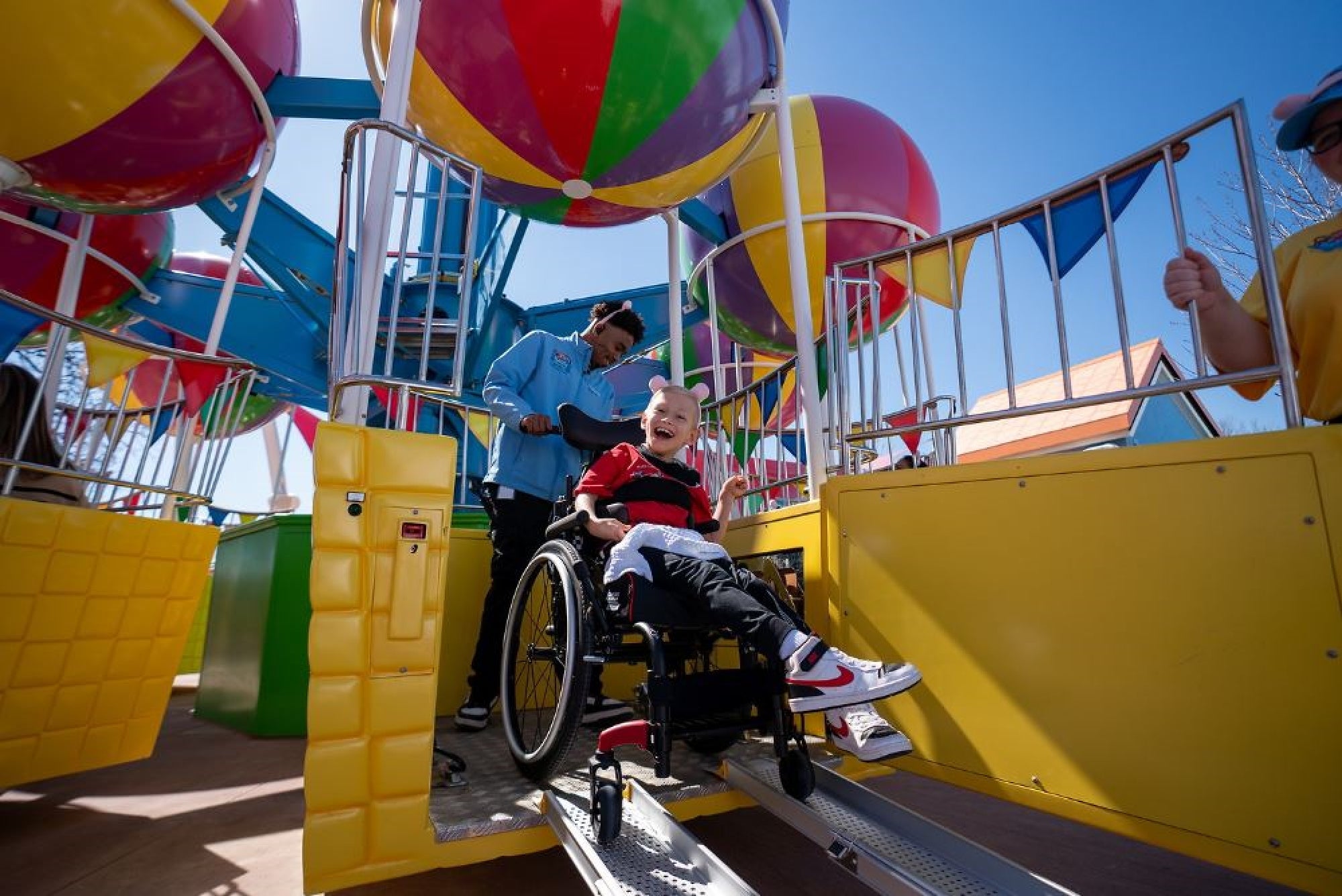Child in a wheelchair smiling while being assisted onto a carnival ride with colorful balloons, accompanied by theme park staff.
