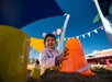 Child smiling and playing in the sand with large orange bucket at a colorful playground under a clear blue sky.