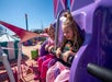 Two children smiling on a purple amusement park ride under a clear blue sky.