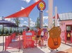 People stand in line at a red amusement park ride under a bright blue sky. A large cartoon cow character appears on a fence nearby.