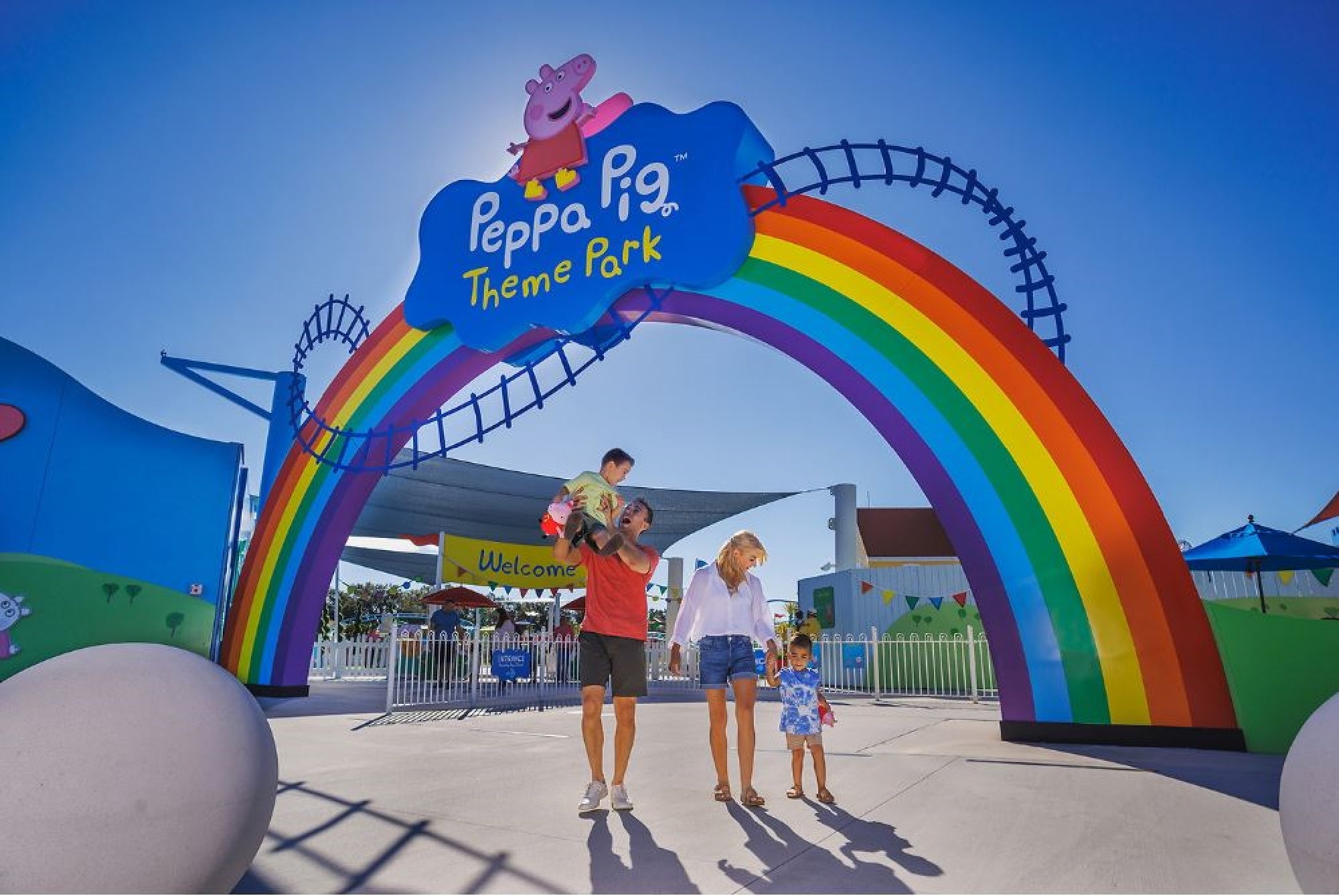 Family of four walking under a rainbow archway at the entrance of Peppa Pig Theme Park on a sunny day.