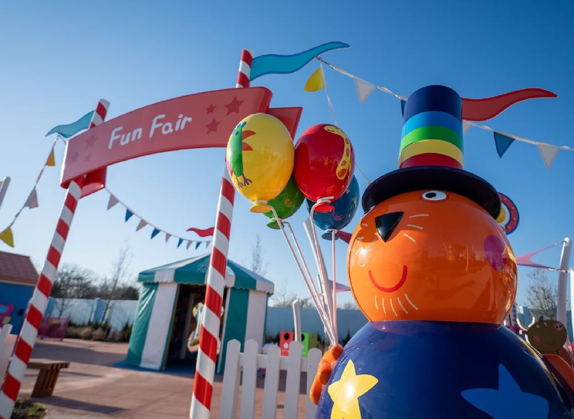 Colorful fun fair entrance with a whimsical orange statue wearing a top hat, holding balloons. Bright blue sky and festive decorations in the background.