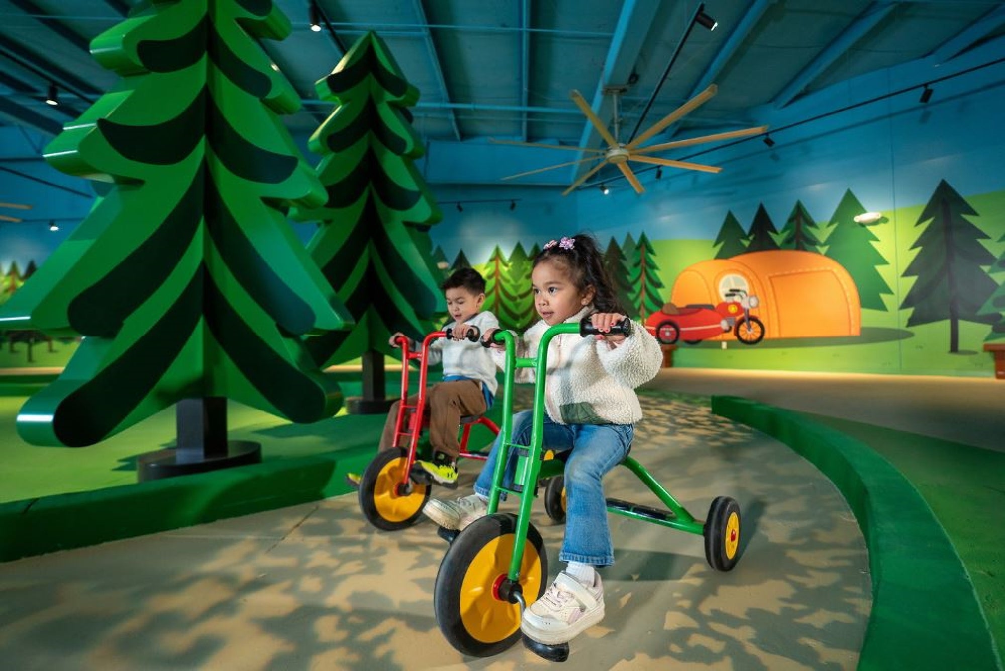 Two children ride tricycles on a path surrounded by painted trees and a tent in a play area.