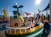 A woman and two children sit smiling in a colorful amusement park boat ride under a clear blue sky.
