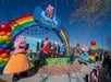 People in Peppa Pig costumes at a colorful outdoor event with balloons, rainbow decor, confetti, and an audience.