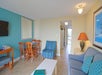 Hotel room with a small dining area, striped armchair, sofa with blue pillows, coffee table, wall art, and a TV. Doorway leads to a hallway and another room.
