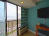 Hotel room with teal walls, a wall-mounted TV, small wooden table, air conditioning unit, and sliding glass door opening to a balcony with an ocean view.