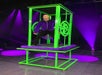 A person stands inside a green and purple illusion box on stage, wearing a purple outfit. Bright purple lights illuminate the scene.