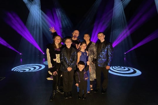 A group of seven people, including children, posing with stage lighting and purple spotlights in the background.