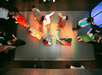 Top view of a group of people dancing on a small square dance floor, illuminated by colorful lights in a dimly lit room.