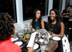 Three women sit at a dining table with menus and drinks, engaged in conversation and smiling at each other.