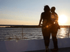 Two people stand side by side on a boat at sunset, gazing at the water with the sun slightly above the horizon.