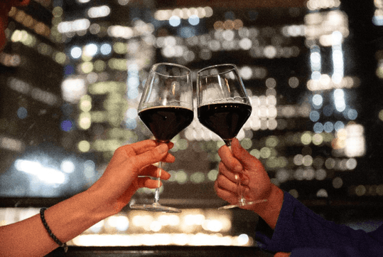 Two hands clinking wine glasses filled with red wine against a blurred cityscape background at night.