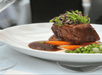 Plate of braised meat topped with microgreens, served with sliced carrots, peas, and a brown sauce.