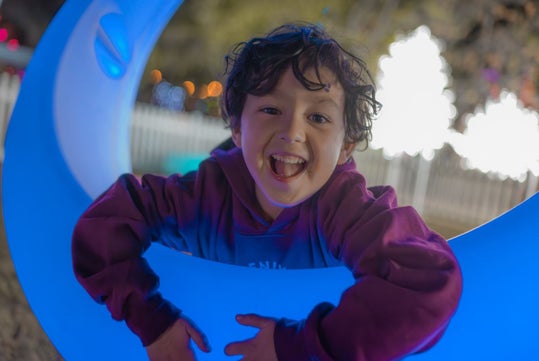 Child smiling and leaning through a blue, circular structure with bright lights in the background.