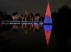 A large conical structure and palm trees decorated with colorful lights are reflected in a calm body of water at night.