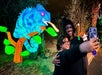 Two people take a selfie in front of a large, illuminated chameleon sculpture at night.