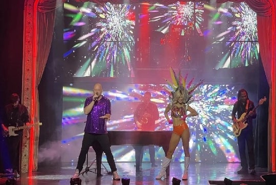 Kyle Martin performing on stage in a purple sequin shirt with a dancer next to him and a band in the background.