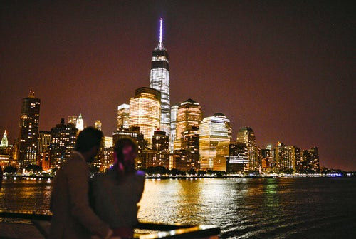Two people stand by a waterfront at night, looking at the illuminated skyline of a city with tall buildings reflecting on the water.