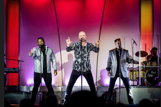 Three men perform energetically on stage with microphones, dressed in stylish jackets, against a vibrant, colorful backdrop. A drummer is visible in the background.