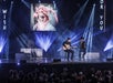Two musicians perform on stage with guitars, seated under spotlights, while a large screen above them displays a person in costume and glasses. An audience watches in the foreground.