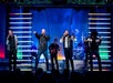 A band of five men performs energetically onstage with colorful lights and a band in the background.