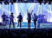 Four performers in shiny jackets sing and dance on a brightly lit stage, backed by a band. An audience watches from below.