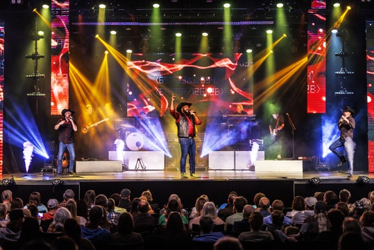 A band performs on a brightly lit stage with colorful spotlights and smoke effects, while a seated audience watches.