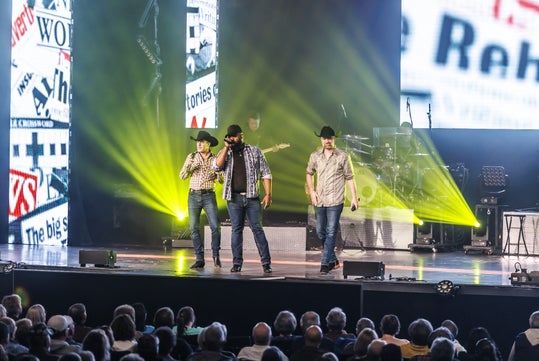 Three men in cowboy hats perform on stage with yellow lights and a news-themed backdrop, in front of an audience.