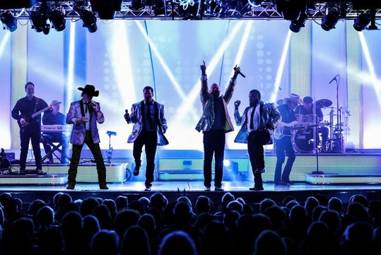 A live band performs energetically on a brightly lit stage with colorful lights. Six musicians are playing instruments and singing, while an audience watches from the foreground.