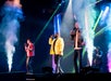 Three male singers perform onstage with microphones, colorful lights, and stage smoke, while musicians play instruments in the background.