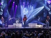 A band performs on stage with dramatic lighting, featuring five musicians and several instruments, as an audience watches from the foreground.